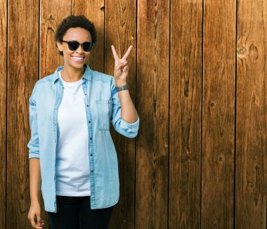 Güzel genç Afro-Amerikan kadın mutlu yüz zafer işareti yaparken kameraya göz kırpıyor ile gülümseyen izole arka plan üzerinde güneş gözlüğü takıyor. İki numaralı.