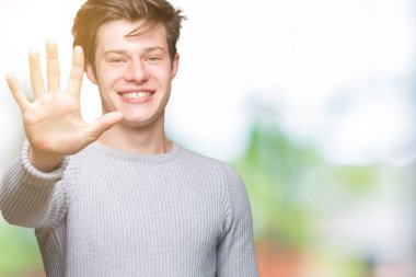 Gösterilen arka plan üzerinde kış kazak giyen genç yakışıklı adam izole ve parmakları ile işaret kendinden emin ve mutlu gülümseyerek süre beş numara.