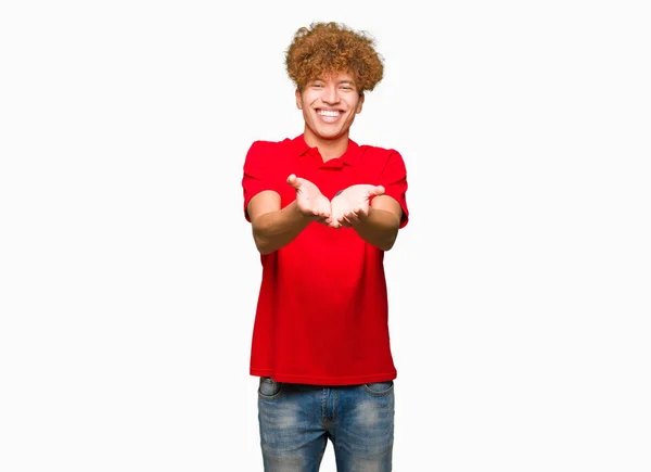 赤いTシャツを着たアフロヘアの若いハンサムな男は 手のひらで微笑み 一緒にジェスチャーを受けたり与えたりします ホールドと保護 — ストック写真