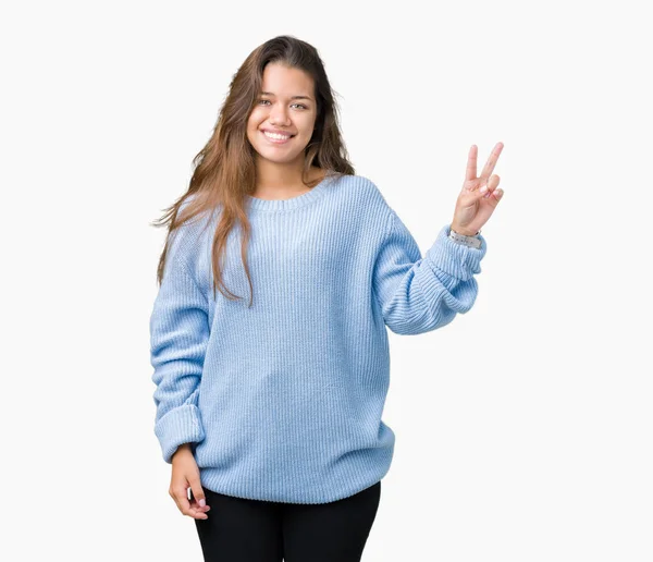 年轻美丽的黑发女人穿着蓝色的冬季毛衣在孤立的背景微笑着与幸福的脸眨眼对着相机做胜利的标志 — 图库照片