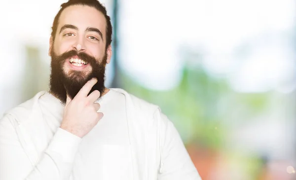 Young man with long hair and beard wearing sporty sweatshirt looking confident at the camera with smile with crossed arms and hand raised on chin. Thinking positive.