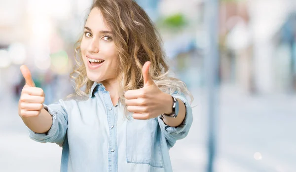 Beautiful young blonde woman over isolated background approving doing positive gesture with hand, thumbs up smiling and happy for success. Looking at the camera, winner gesture.