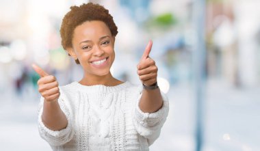 El ile pozitif jest yapıyor onaylama izole arka plan üzerinde kazak giyen güzel genç Afro-Amerikan kadın başparmak gülümseyen ve başarı için mutlu. Kamera, kazanan jest arıyorsunuz.