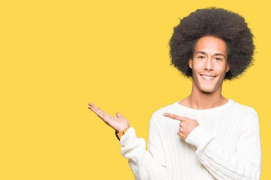 Afro saçlı giyen kış kazak hayret ve el ile sunulması ve parmak ile işaret ederken kameraya gülümseyen genç Afro-Amerikan adam.