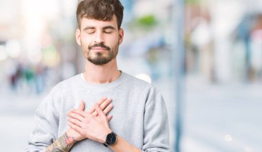 Yakışıklı delikanlı ile gülümseyen izole arka plan üzerinde sweatshirt giyen kapalı gözler ve yüzünde minnettar jest ile göğüs üzerinde eller. Sağlık kavramı.