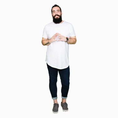 Genç hippi erkek uzun saç ve sakal casual beyaz t-shirt eller birlikte giyen ve parmakları ile rahat ve neşeli gülümseyen geçti. Başarı ve iyimser