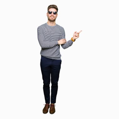 Kameraya bakarak yan el ve parmak ile işaret eden genç yakışıklı adam, yüzünde kocaman bir gülümseme ile güneş gözlüğü takıyor.