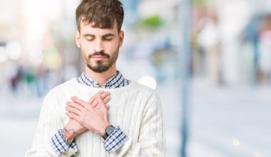 Yakışıklı delikanlı ile gülümseyen izole arka plan üzerinde kış kazak giyen kapalı gözler ve yüzünde minnettar jest ile göğüs üzerinde eller. Sağlık kavramı.