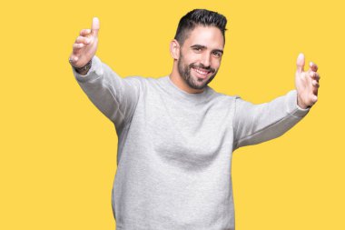Sarılmak için kollarını açarak gülümseyerek kameraya bakarak izole arka plan üzerinde sweatshirt giyen genç yakışıklı adam. Neşeli ifade kucaklayan mutluluk.