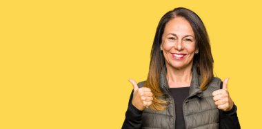 Güzel orta yaş kadın giyen kış yelek başarı işareti el ile olumlu jest yapıyor, gülümseyen ve mutlu kadar başparmak. Neşeli ifade ile kamera bakarak, kazanan jest.