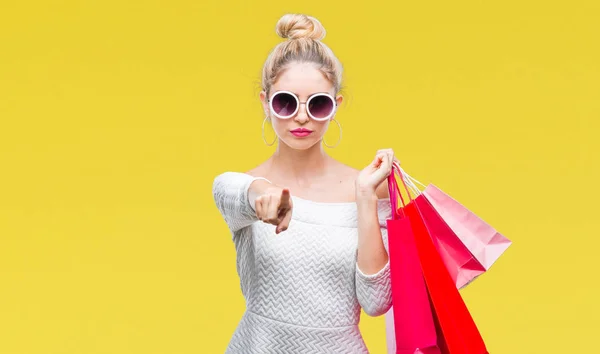 若い金髪美女持株ショッピング バッグ カメラと指で指している孤立した背景に 手に署名 正面からポジティブで自信のジェスチャー — ストック写真