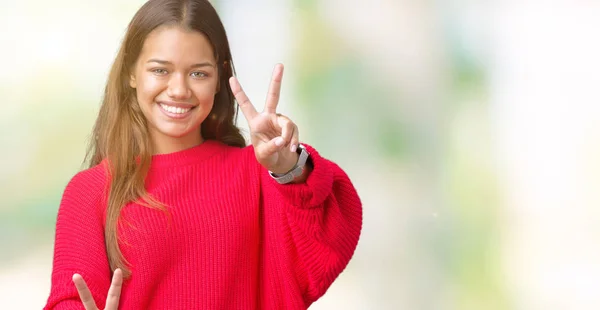 年轻美丽的黑发女人穿着红色的冬季毛衣在孤立的背景微笑着看着相机显示手指做胜利的标志 — 图库照片