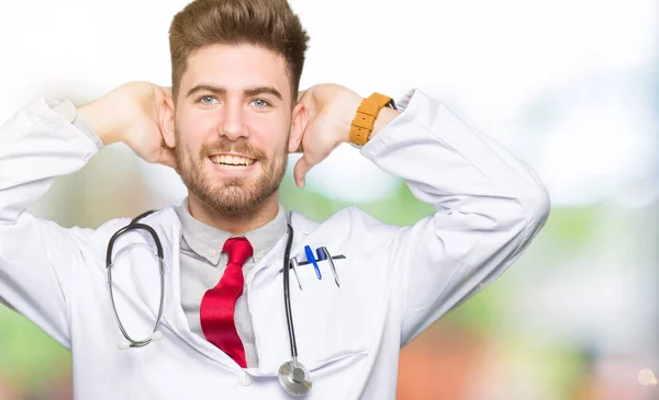 年轻英俊的医生男子穿着医疗外套放松和伸展与手臂和双手背后的头部和颈部 面带微笑的快乐 — 图库照片