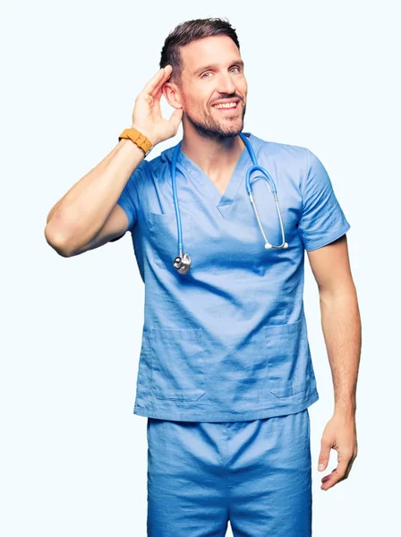 英俊的医生穿医疗制服在孤立的背景微笑与手在耳朵听谣言或流言蜚语 耳聋概念 — 图库照片