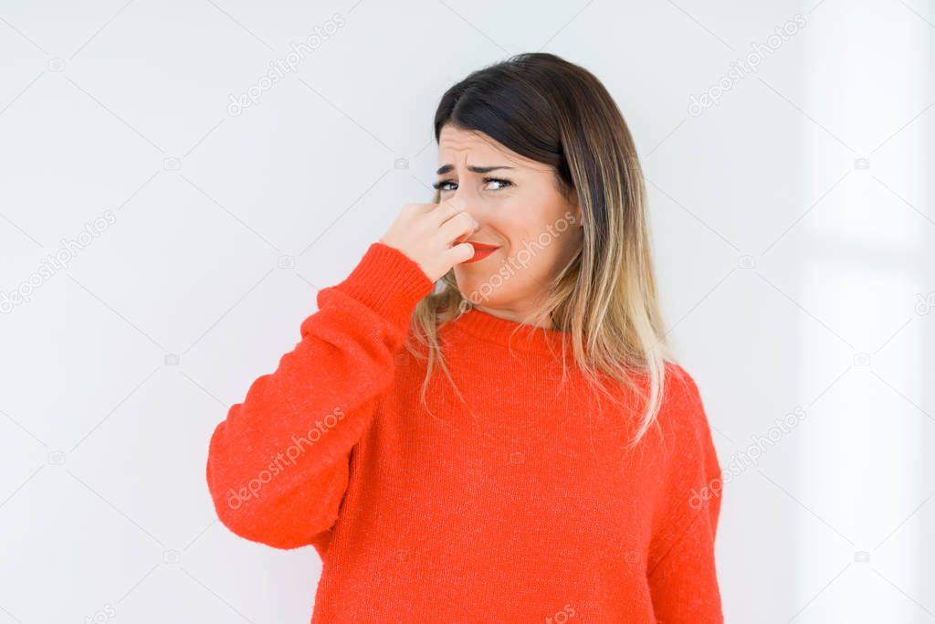 Young woman wearing casual red sweater over isolated background smelling something stinky and disgusting, intolerable smell, holding breath with fingers on nose. Bad smells concept.