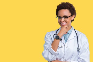 Genç Afro-Amerikan doktor kadın emin çapraz kollar ve çeneme kaldırdı el ile gülümseme ile kameraya bakarak izole arka plan üzerinde tıbbi bir palto giyiyor. Pozitif düşünce.