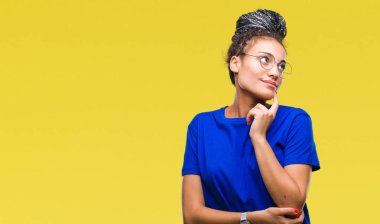 Genç saç Afro-Amerikan Kız çene düşünme hakkında soru, dalgın ifade üzerine el ile izole arka plan üzerinde gözlük takan örgülü. Düşünceli yüzüyle gülümsüyor. Şüphe kavramı.