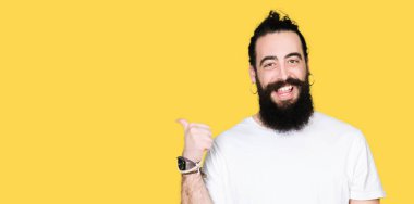 Uzun saçlı ve sakal ile genç hipster adam rahat beyaz t-shirt mutlu yüz görünümlü ve başparmak yukarı ile yan işaret gülümseyen giyiyor.