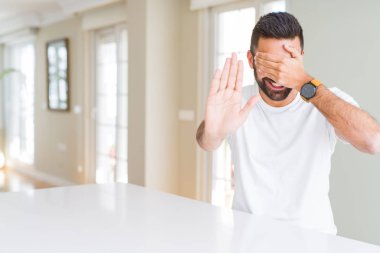 Yakışıklı İspanyol adam evde rahat beyaz t-shirt elleri ile gözleri kaplayan ve üzgün ve korku ifade ile stop jest yapıyor. Utanmış ve olumsuz kavram.