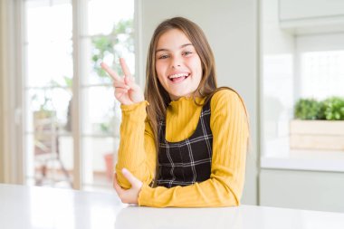 Young beautiful blonde kid girl wearing casual yellow sweater at home smiling with happy face winking at the camera doing victory sign with fingers. Number two.