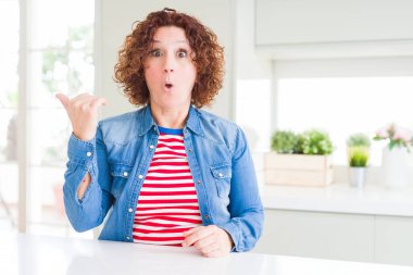Evde kot ceket giyen kıvırcık saçlı orta yaş yaşlı kadın yan el parmağı ile işaret şaşırttı, açık ağız şaşkın ifade.