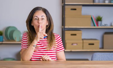 Orta yaş yaşlı kadın evde masada oturan dudaklarında parmak ile sessiz olmak isteyen. Sessizlik ve gizli kavram.