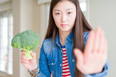 Güzel Asyalı kadın ciddi ve kendinden emin ifade, savunma jesti ile durdurmak işareti yapıyor açık el ile yeşil taze brokoli yeme
