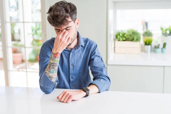 Young man wearing casual shirt sitting on white table tired rubbing nose and eyes feeling fatigue and headache. Stress and frustration concept.