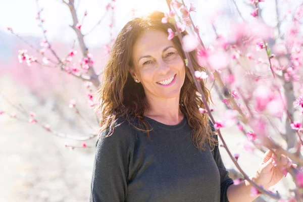 Beautiful middle age woman in the middle of pink peach flowers and trees smiling cheerful, colorful sunny day of spring