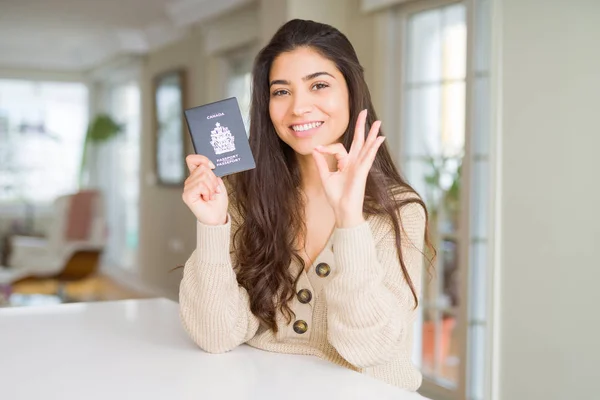 Young woman holding passport of Canada doing ok sign with fingers, excellent symbol