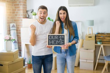 Young beautiful couple holding blackboard with our first home text at new house screaming proud and celebrating victory and success very excited, cheering emotion