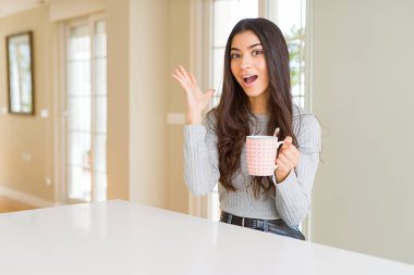 Bir fincan kahve içen genç kadın çok mutlu ve heyecanlı, kazanan yüz ifadesi zaferi büyük bir gülümsemeyle kutluyor ve ellerini kaldırıyor.