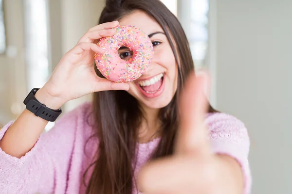 美丽的年轻女子吃粉红色的巧克力片甜甜圈快乐与大微笑做Ok标志 拇指用手指 优秀的标志 — 图库照片