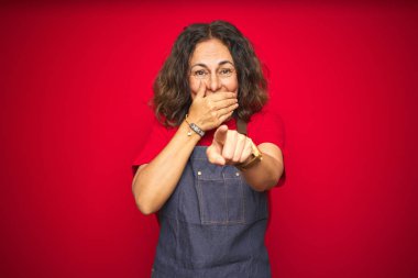 Kırmızı izole arka plan üzerinde önlük üniforma giyen orta yaş yaşlı kadın size gülüyor, ağız üzerinde el ile kameraya parmak işaret, utanç ifadesi