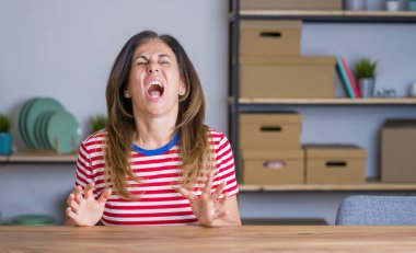 Orta yaş yaşlı kadın evde masada oturan kızgın ve deli çığlık sinirli ve öfkeli, öfke ile bağırarak. Öfke ve agresif kavram.