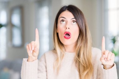 Evde kış kazak giyen genç güzel kadın hayrete ve yukarıya bakıyor ve parmakları ile işaret ve silah kaldırdı şaşırttı.