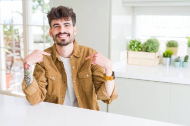 Young man wearing casual jacket sitting on white table looking confident with smile on face, pointing oneself with fingers proud and happy.