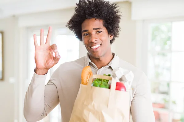 African American man holding groceries bag with fresh vegetables at home doing ok sign with fingers, excellent symbol