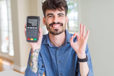 Young man holding dataphone point of sale as payment doing ok sign with fingers, excellent symbol