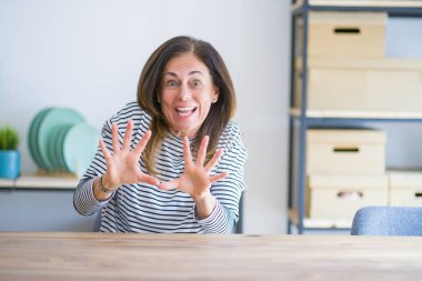 Orta yaş yaşlı kadın evde masada oturan korkmuş ve korku ile korkmuş ifade elleriyle jest durdurmak, şok içinde bağırarak. Panik kavramı.