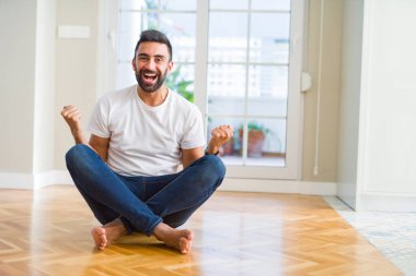 Yakışıklı İspanyol adam evde yerde oturan rahat t-shirt giyen çok mutlu ve heyecanlı kolları kaldırdı ile kazanan jest yapıyor, gülümseyerek ve başarı için çığlık. Kutlama konsepti.