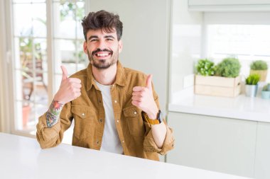 Young man wearing casual jacket sitting on white table success sign doing positive gesture with hand, thumbs up smiling and happy. Looking at the camera with cheerful expression, winner gesture.