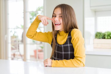 Young beautiful blonde kid girl wearing casual yellow sweater at home Pointing with hand finger to face and nose, smiling cheerful. Beauty concept