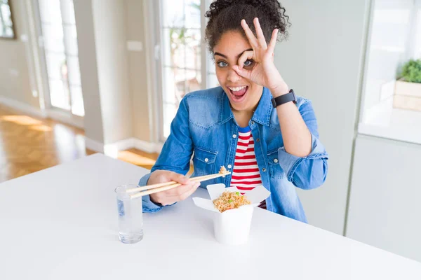 Young african american woman eating asian noodles from delivery box with happy face smiling doing ok sign with hand on eye looking through fingers
