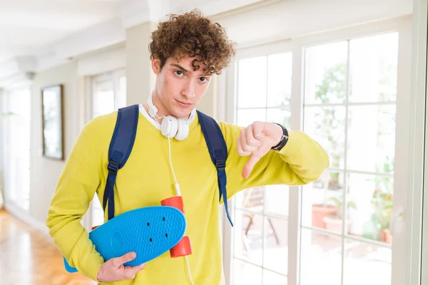 怒った顔でスケートボードを保持ヘッドフォンやバックパックを身に着けている若い学生の男 親指ダウンで嫌いを示す負の記号 拒絶概念 — ストック写真