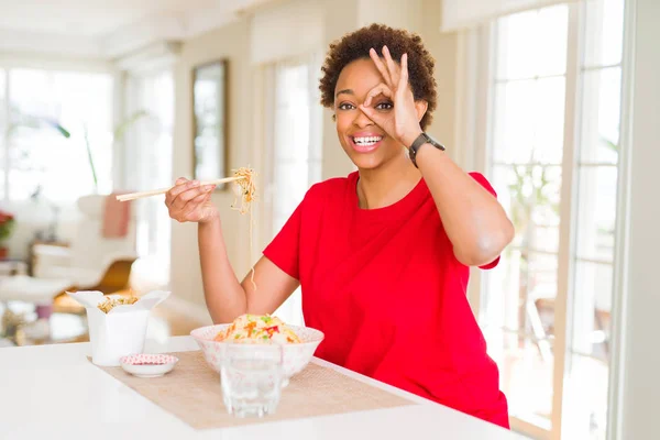 Young african american woman with afro hair eating asian food at home with happy face smiling doing ok sign with hand on eye looking through fingers