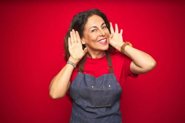 Orta yaş yaşlı kadın kırmızı izole arka plan üzerinde önlük üniforma giyen kulak jesti iki elini duymak için çalışıyorum, dedikodu için meraklı. İşitme sorunu, sağır