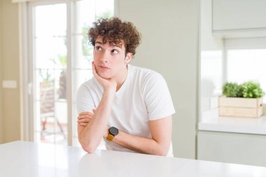 Evde eli çenesinde beyaz tişört giyen genç yakışıklı bir adam soru sormayı düşünüyor, düşünceli bir ifade. Düşünceli bir yüzle gülümsüyor. Şüphe kavramı.