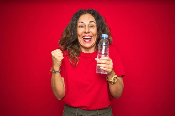 Middle age senior woman holding plastic water bottle over red isolated background screaming proud and celebrating victory and success very excited, cheering emotion