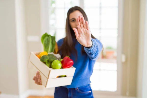 Beautiful young woman holding wooden box full of healthy groceries with open hand doing stop sign with serious and confident expression, defense gesture
